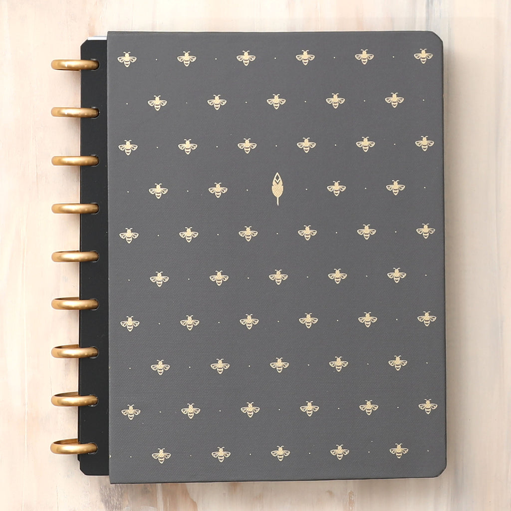 A hard cover featuring black background and busy bee design for this inkwell press 7x9 planner system featuring gold discs.
